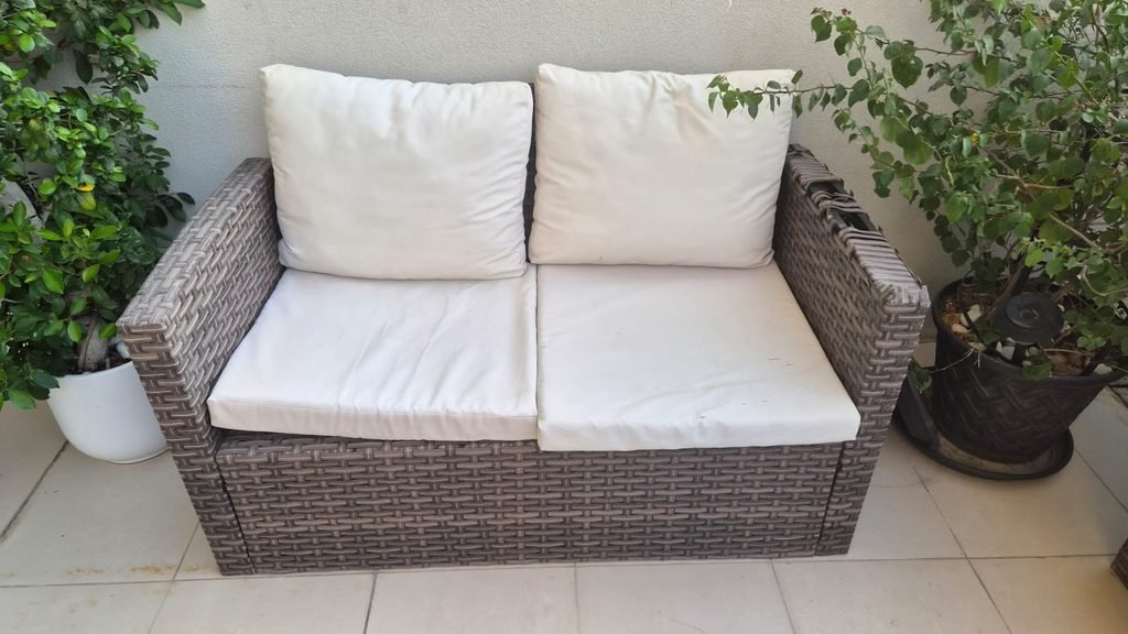 A brown wicker-style three-seater outdoor sofa with white cushions. This image can support content about bulky sofa removal, outdoor furniture pickup, and junk removal services in Abu Dhabi.