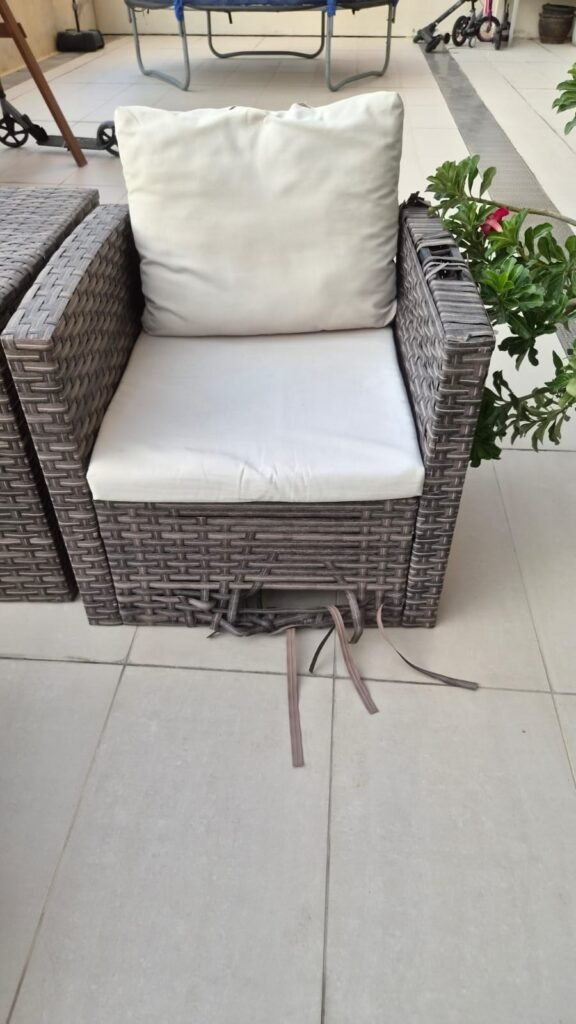 An outdoor wicker armchair with white seat and back cushions and damaged weaving near the base. The image can support content about old furniture pickup and removal services.